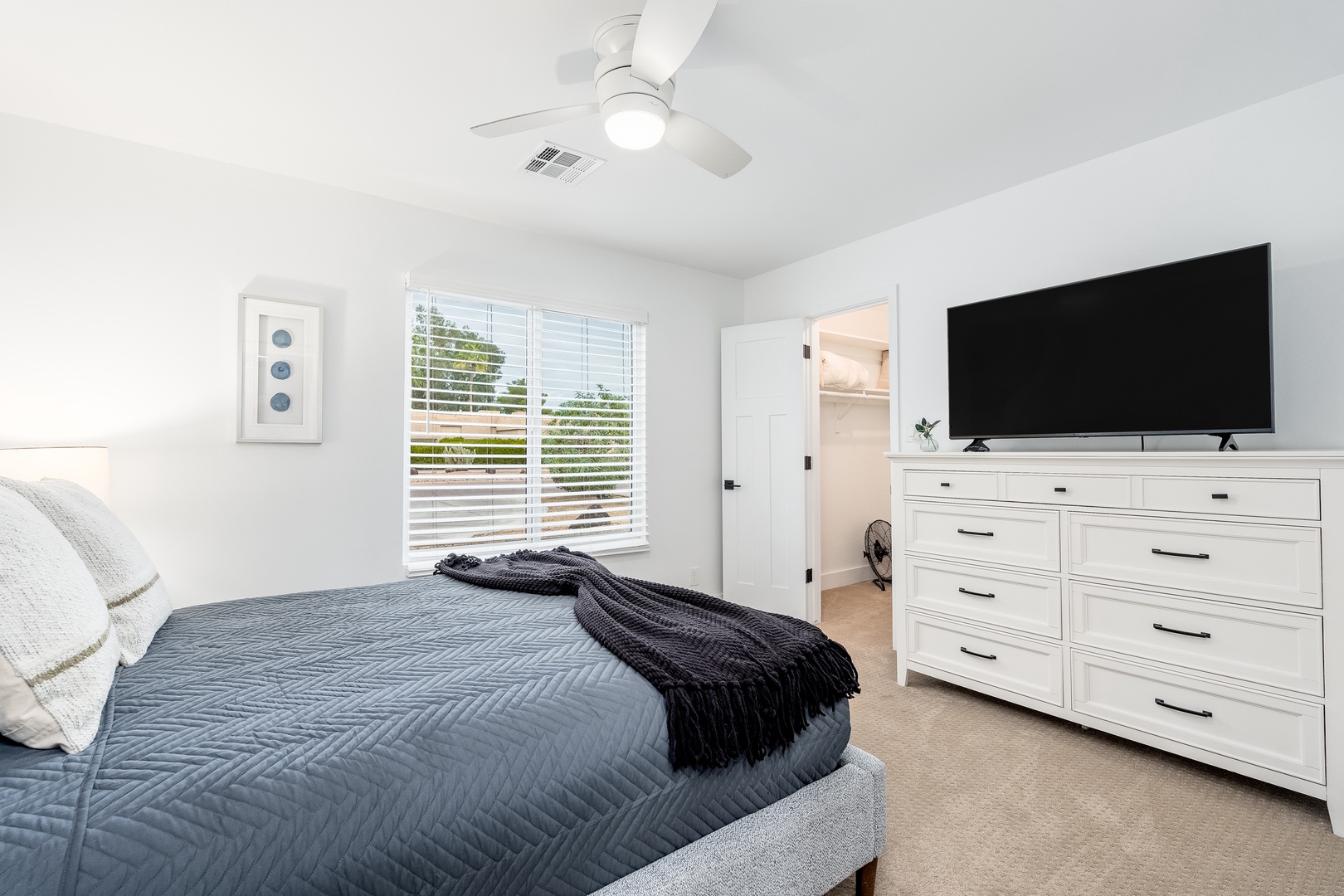 Unwind in your serene bedroom featuring a plush bed, ample storage in the white dresser, a mounted smart TV for entertainment, and a ceiling fan for your comfort.
