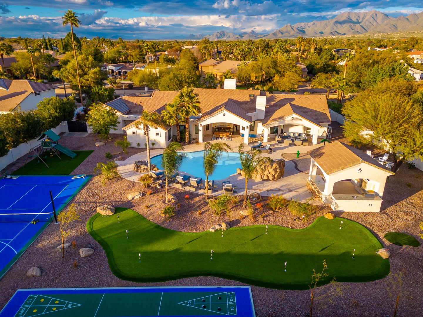 Aerial view showcasing a luxury desert property with a private swimming pool, putting green, sport court, and covered patio areas, set against a backdrop of majestic mountains and a tree-lined neighborhood.