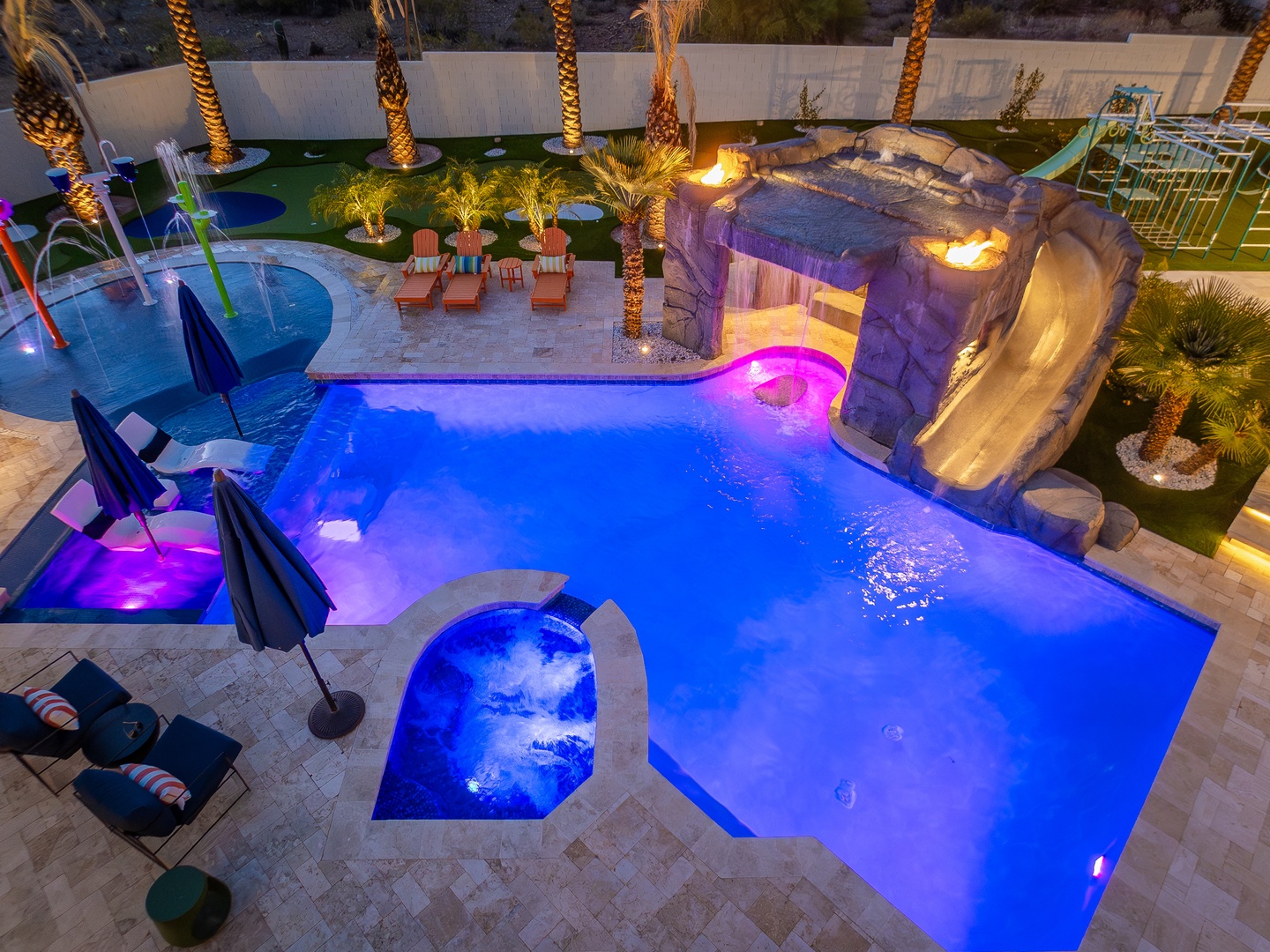 This resort-style backyard offers an illuminated pool with water slide, hot tub, palm trees, and lounge chairs—your private oasis for evening relaxation and fun.