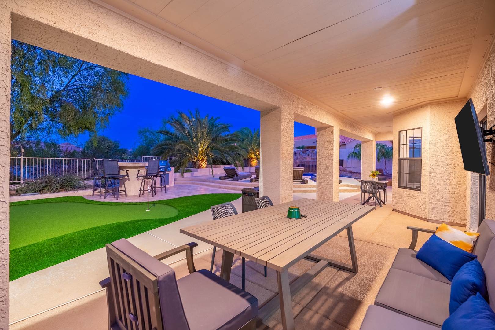 Evening falls over your private covered patio, where outdoor dining and putting green await beneath a vibrant desert sky with palms swaying in the distance.