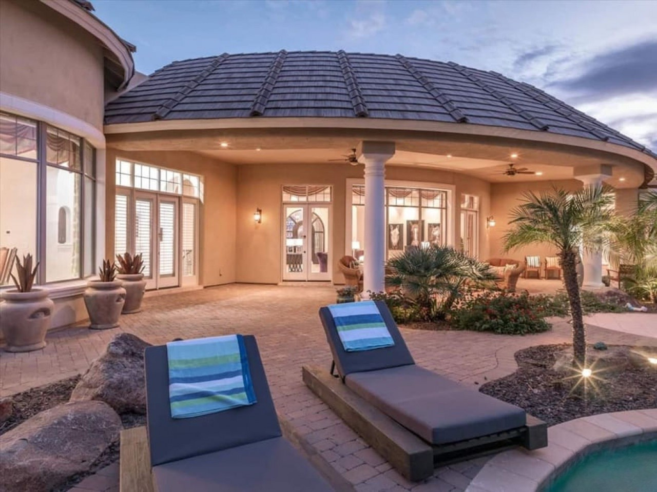 Unwind by the pool on cushioned loungers framed by swaying palms and desert landscaping. The covered patio invites evening relaxation with soft lighting and elegant architectural details at dusk.