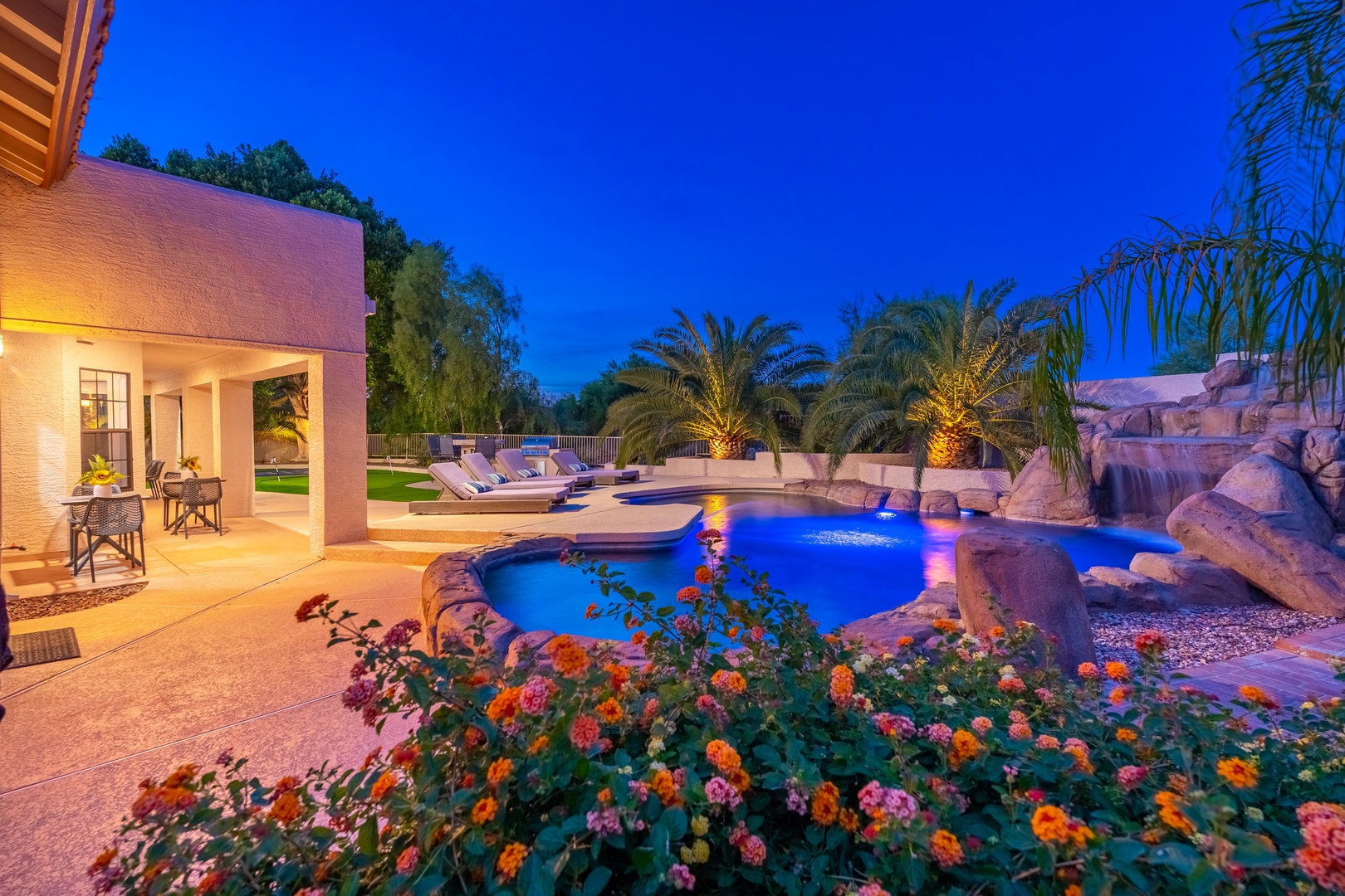 Backyard oasis features an illuminated pool with natural rock waterfall, tropical landscaping, covered patio, and vibrant desert blooms—a stunning evening retreat under open skies.