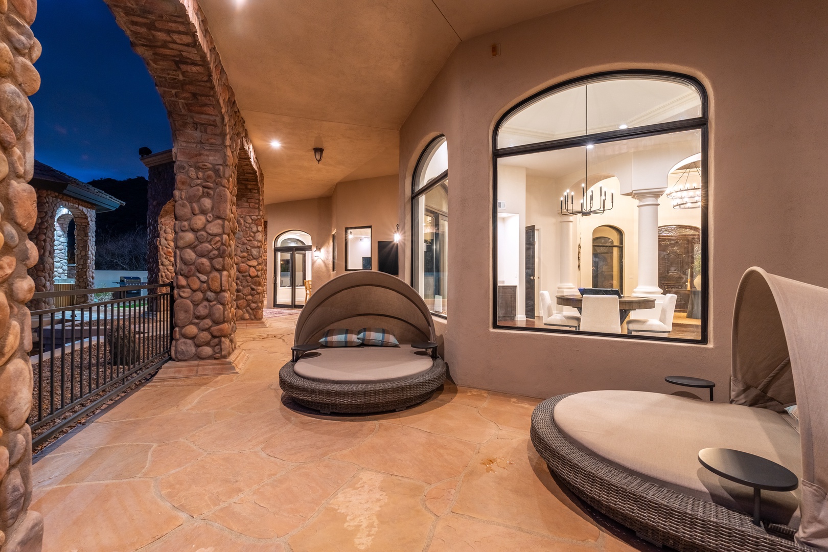Luxurious covered patio with stone accents and modern daybed loungers for evening relaxation under ambient lighting.