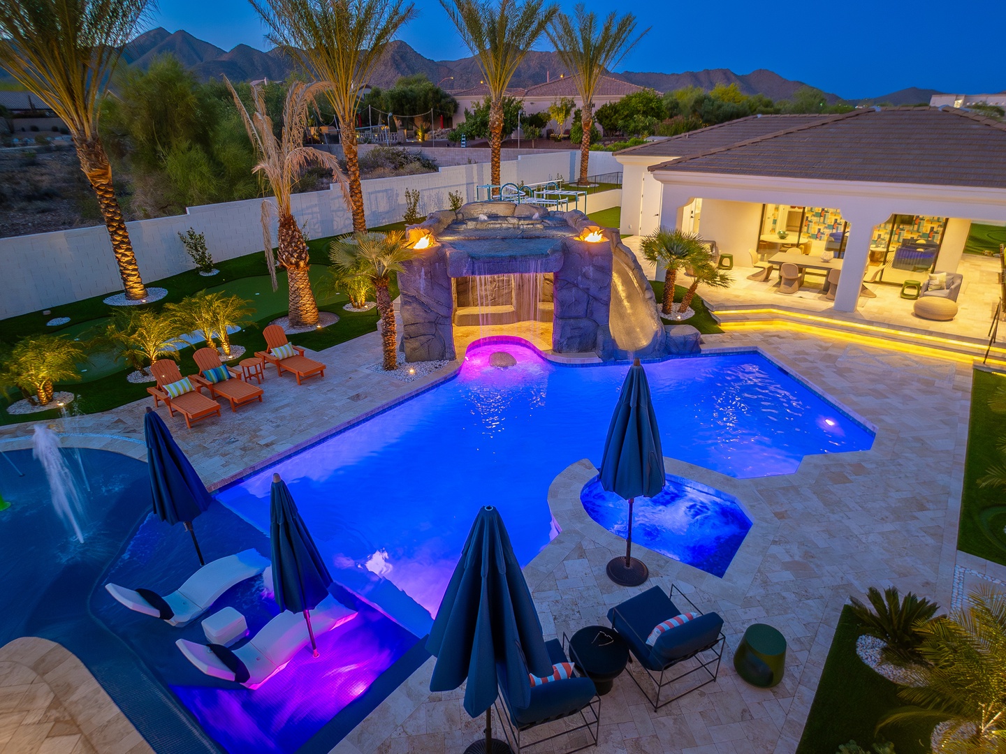Stunning desert oasis with illuminated pool, waterfall grotto, and mountain backdrop at twilight—your private paradise awaits.