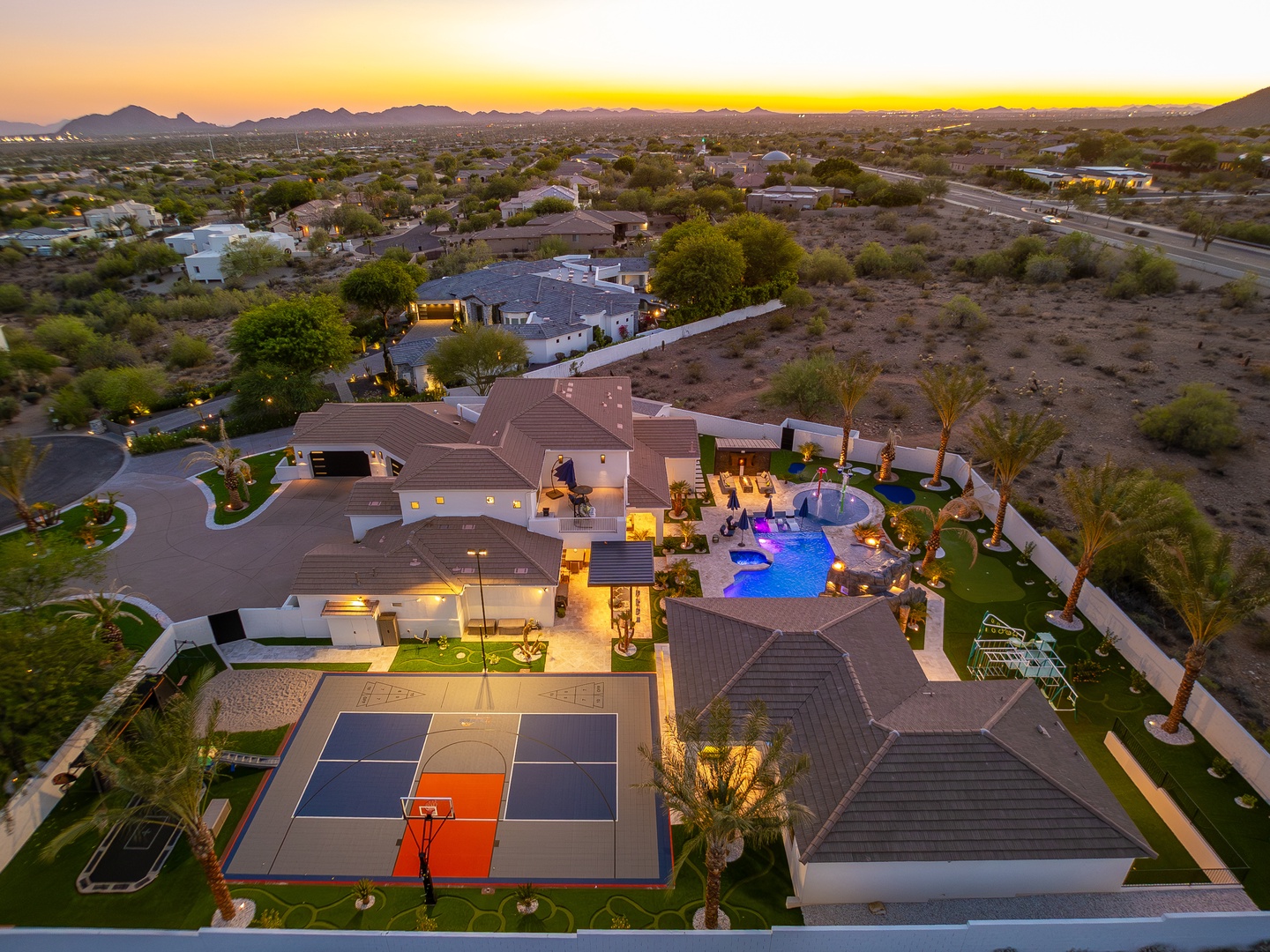 Luxury desert estate with mountain backdrop, outdoor basketball court, resort-style pool, and sprawling desert landscape at sunset.