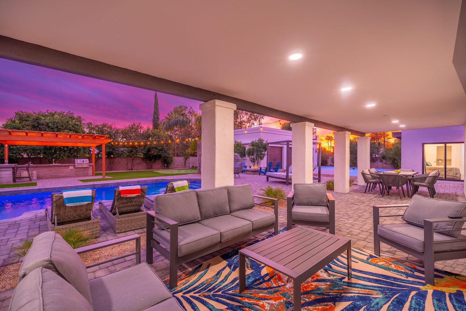 A stunning sunset paints the sky as the property displays covered outdoor seating, poolside relaxation areas, and multiple entertaining spaces across a beautifully landscaped backyard retreat.