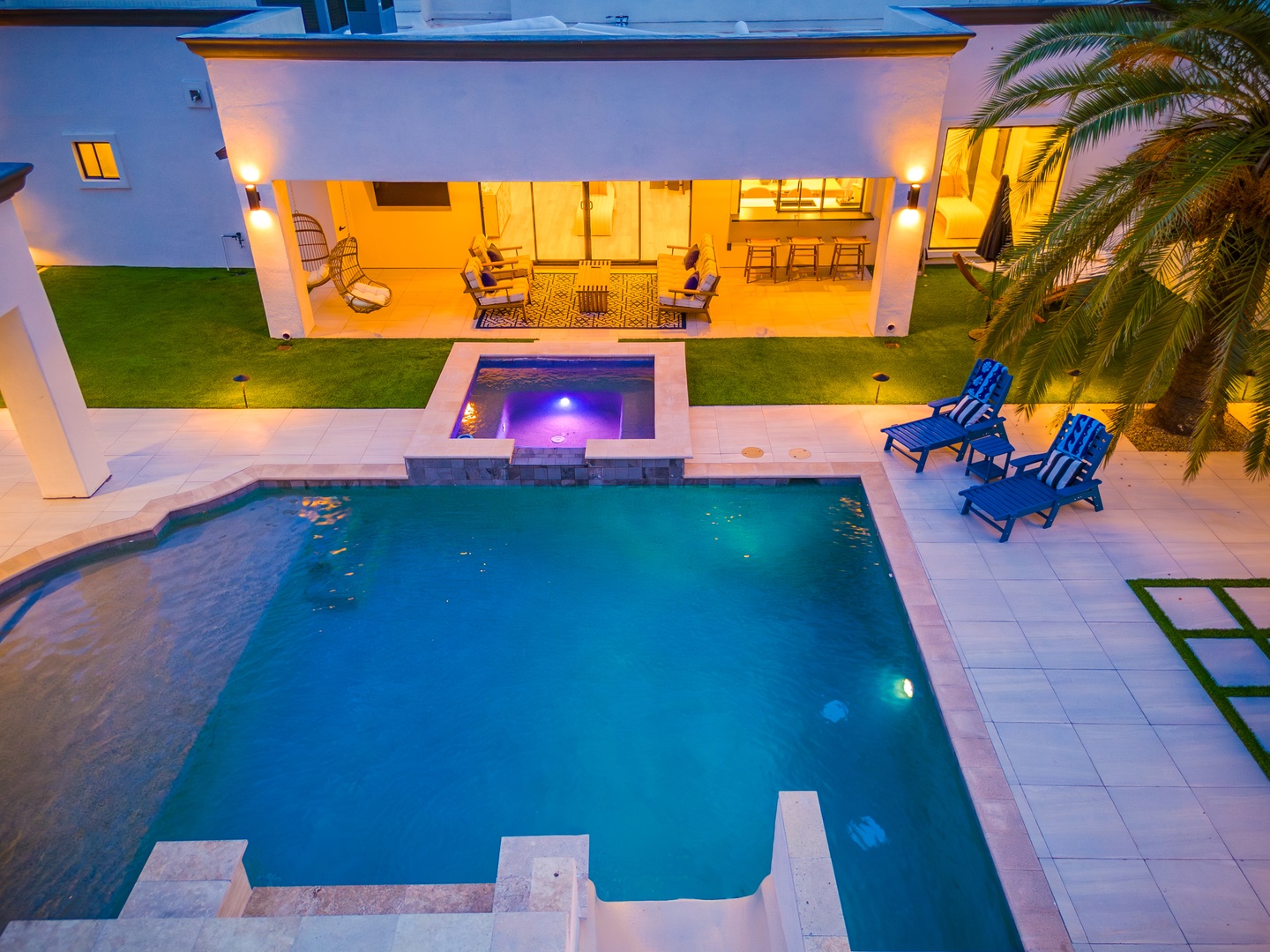 This aerial view showcases the property's lush pool and hot tub oasis framed by palm trees, with a covered patio featuring inviting lounge seating and a cozy atmosphere at dusk.