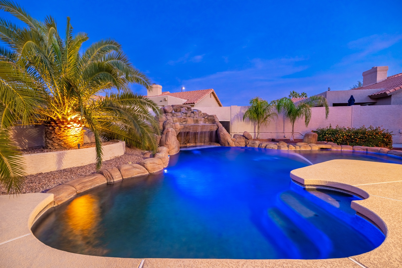 A stunning resort-style pool glows under ambient lighting, featuring a cascading stone waterfall, inviting spa, and tropical palm trees—your private desert oasis awaits.