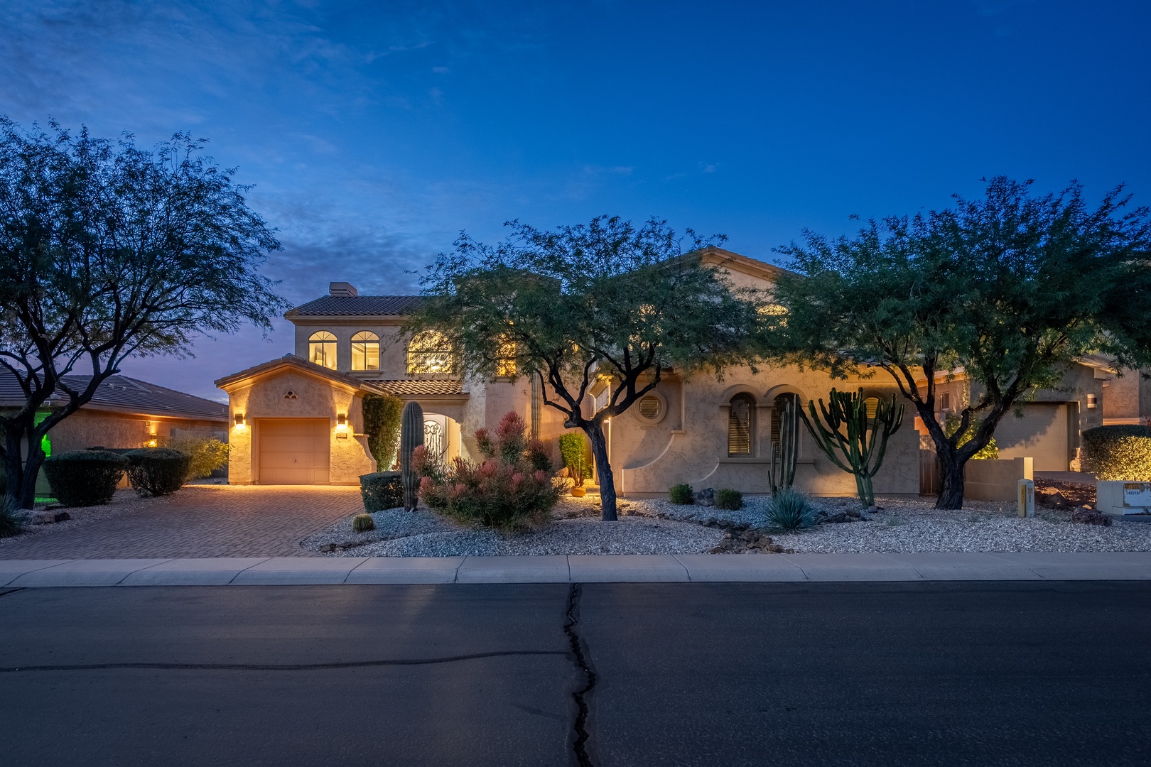 Twilight falls on this Southwestern-style home, featuring stucco walls, arched windows, and desert landscaping framed by native trees and ambient lighting.