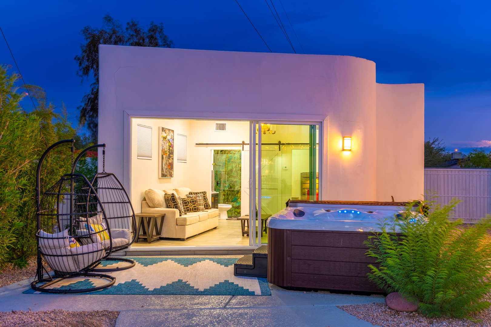 A modern desert retreat features a private hot tub, cozy outdoor seating, and glass doors opening to a stylish living space—ideal for unwinding under vibrant evening skies.