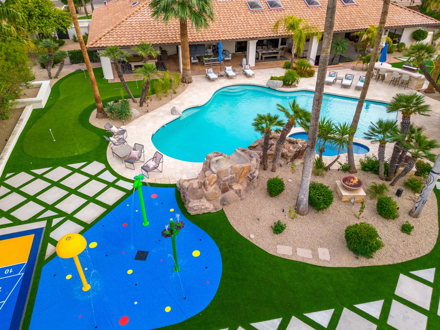 Aerial view of the property showcasing a sparkling pool, minigolf, and a splash pad surrounded by palm trees, lush landscaping, and covered outdoor seating areas under a terrace.
