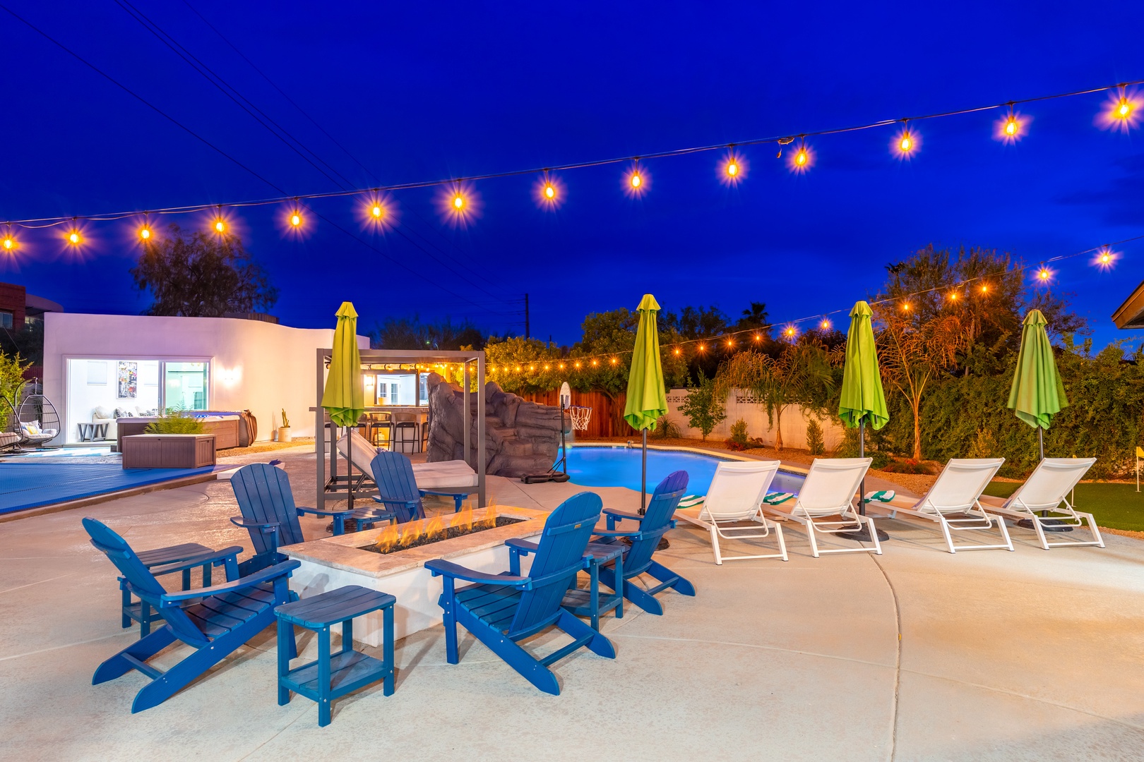 Stunning evening ambiance awaits in this expansive outdoor entertainment area, complete with a sparkling pool, cozy fire pit, hot tub, and festive string lights overhead—your perfect desert retreat.