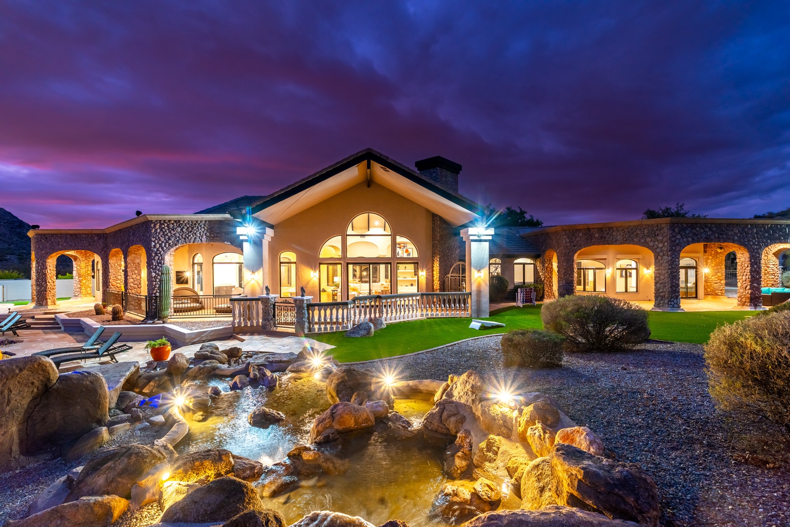 Stunning property facade illuminated at twilight, showcasing beautiful stone architecture, elegant arched windows, and serene water feature with ambient lighting throughout the landscaped grounds.