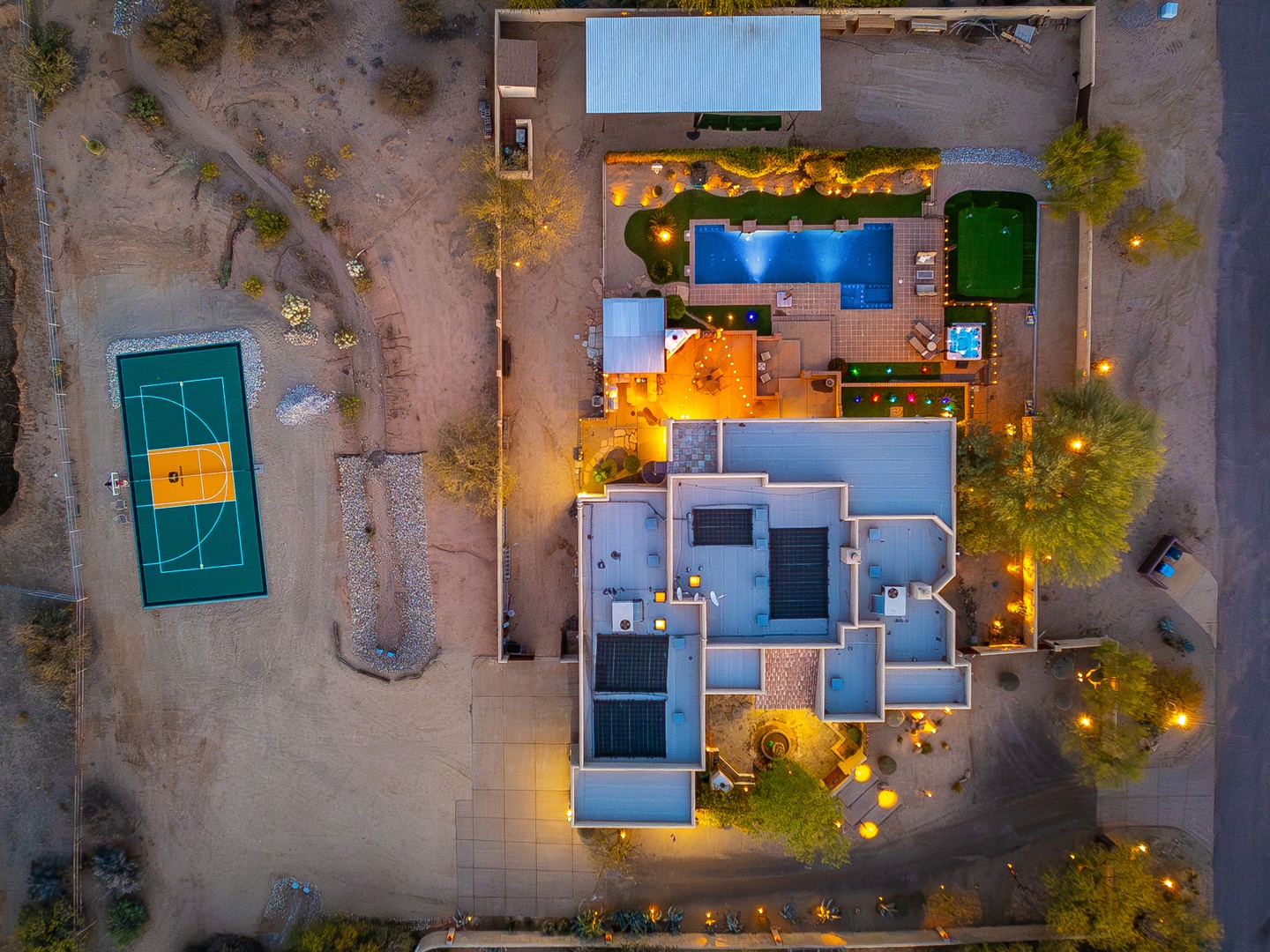 Aerial view of the property at dusk, showcasing the main building, swimming pool, sports court, and beautifully lit outdoor spaces set against the desert landscape.