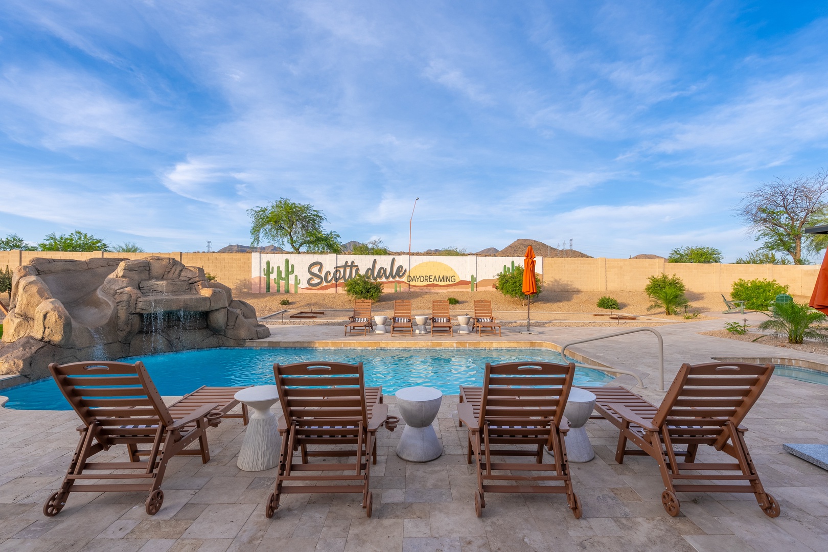 Poolside loungers overlook turquoise waters and a cascading rock waterfall, with desert mountains framing the horizon—perfect for sunny days of relaxation.