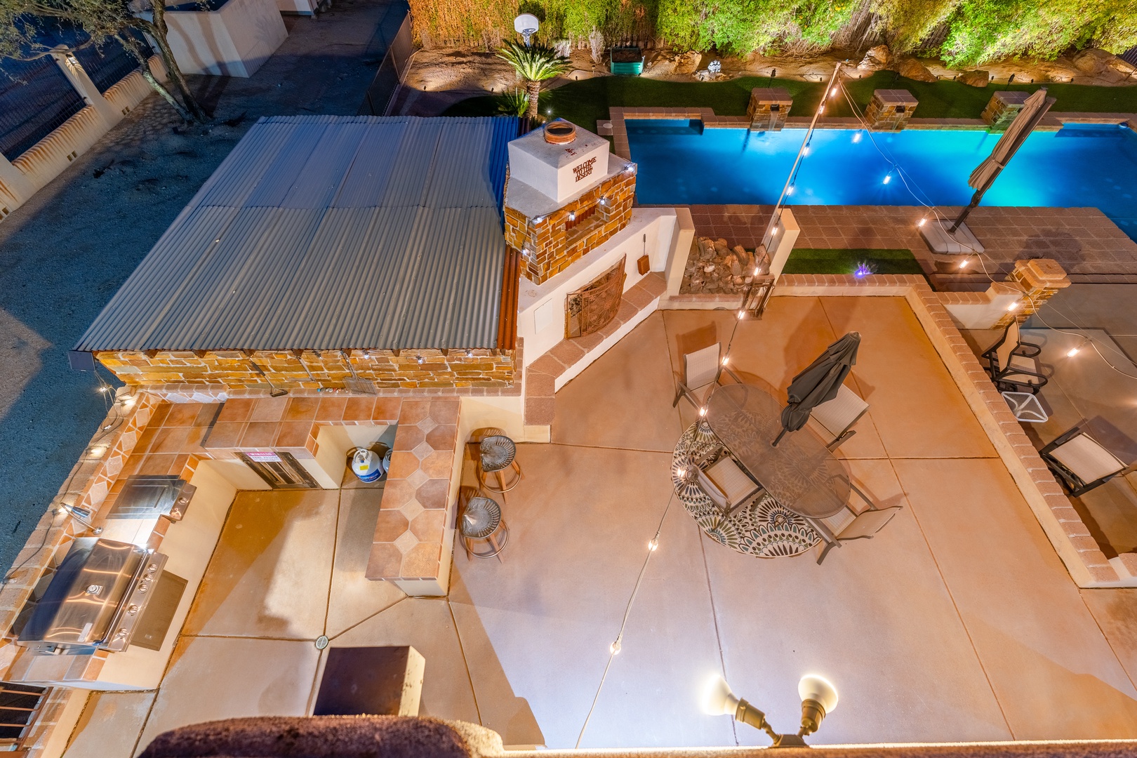 Aerial view showcasing a beautifully landscaped outdoor entertainment area with pool, integrated BBQ facility, covered dining spaces, and surrounding grounds illuminated at night.