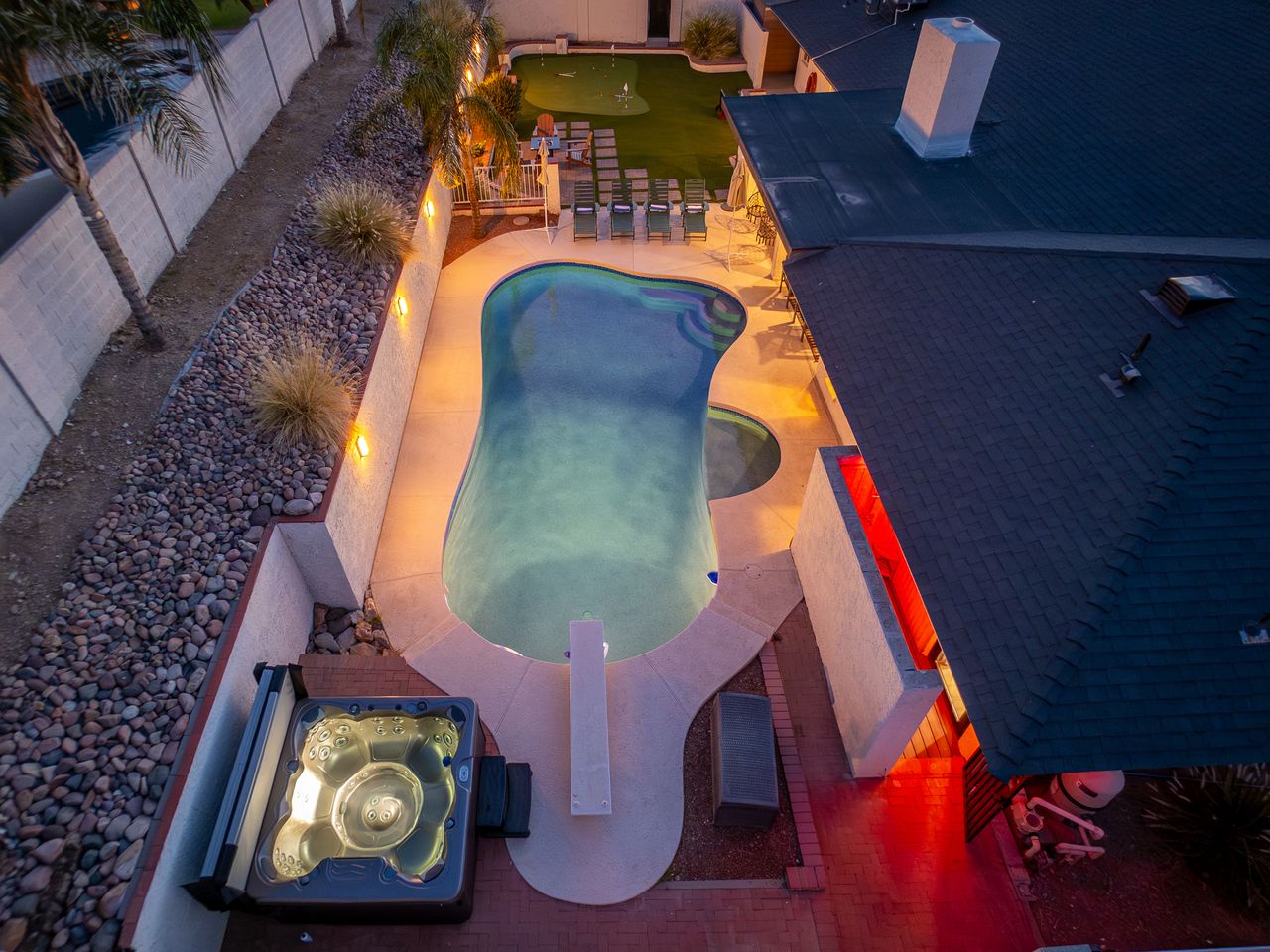 An aerial view captures the property's illuminated outdoor entertainment area at dusk, featuring a freeform pool, spa, artificial turf lounge, and paver surrounds with ambient lighting.