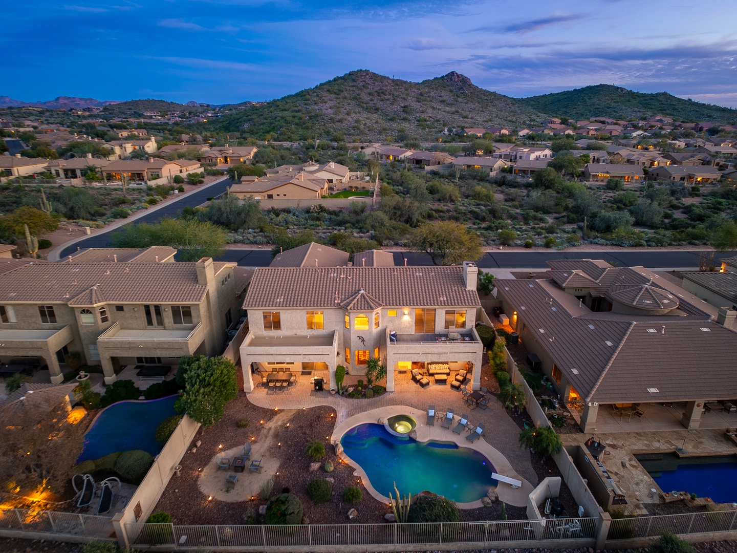 Aerial view of a luxury desert vacation home with private pool and mountain backdrop, situated in a peaceful residential neighborhood at dusk.