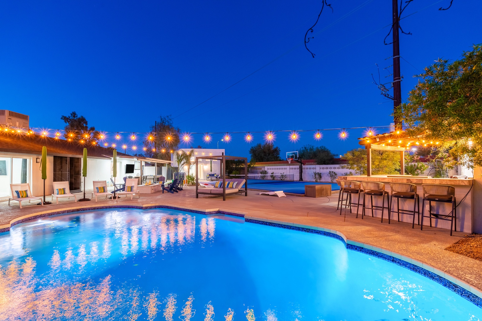 A glowing oasis awaits under string lights, with dual pools, poolside seating, an outdoor bar, and lush landscaping—perfect for evening relaxation and entertaining.