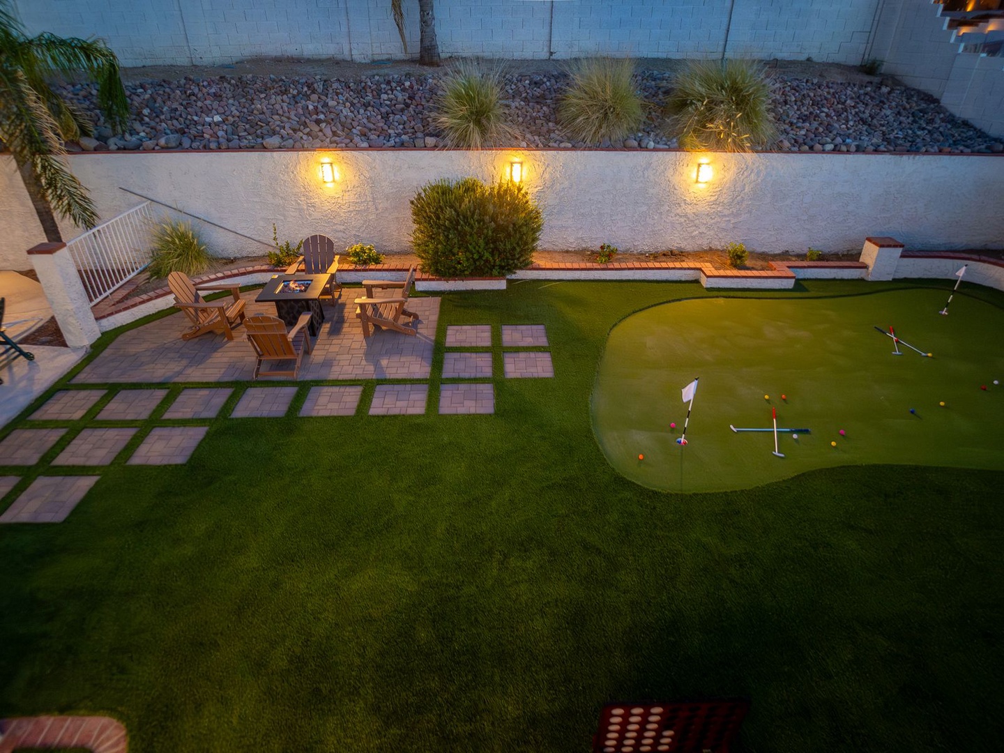 An elevated view showcases the property's private backyard featuring a custom putting green, wooden seating area with fire pit, decorative pathway pavers, and lush landscaping illuminated by warm wall lighting.