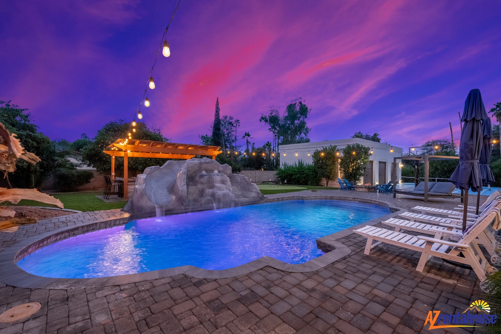 Stunning outdoor pool area featuring dramatic purple sunset skies, string lighting, rock waterfall, and comfortable loungers for evening relaxation.