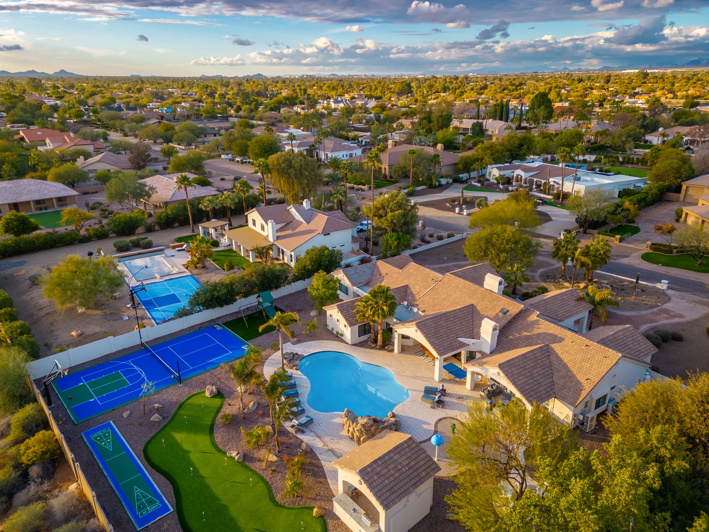 Aerial view of a luxury vacation rental situated in a tranquil residential neighborhood, featuring private pool, tennis court, and putting green surrounded by palm trees and mountain vistas.