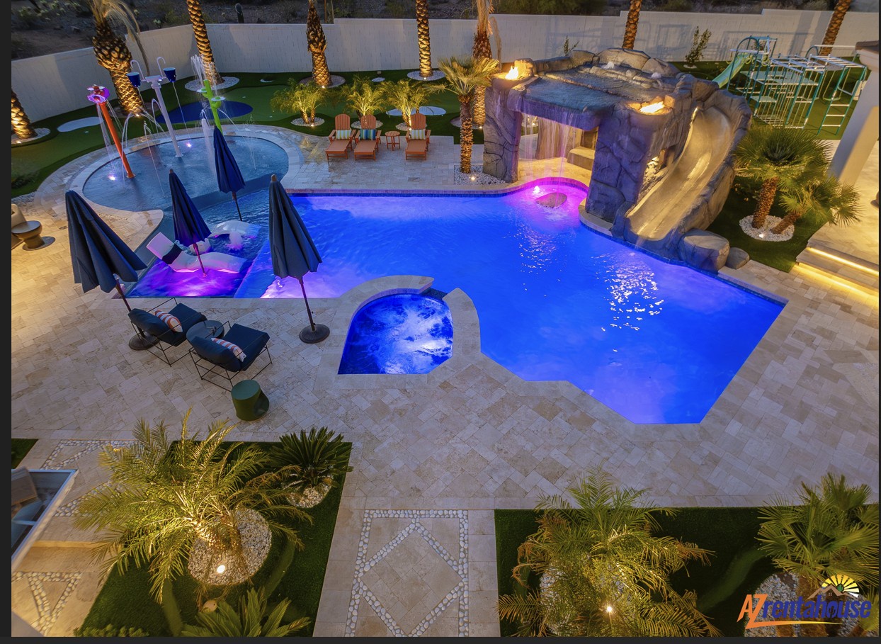 Stunning aerial view of the illuminated pool area featuring a water slide, hot tub, and tropical landscaping with palm trees.