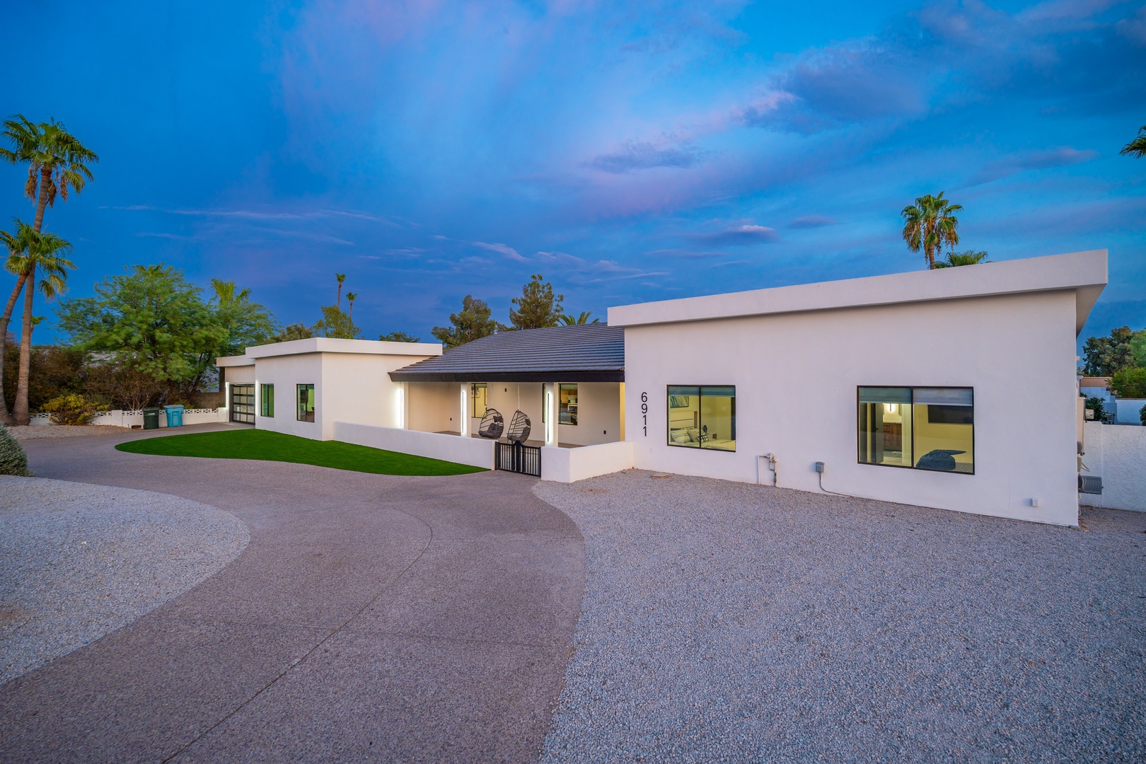 Modern desert retreat framed by palm trees and mountain views, with spacious driveway and manicured lawn welcoming guests at twilight.