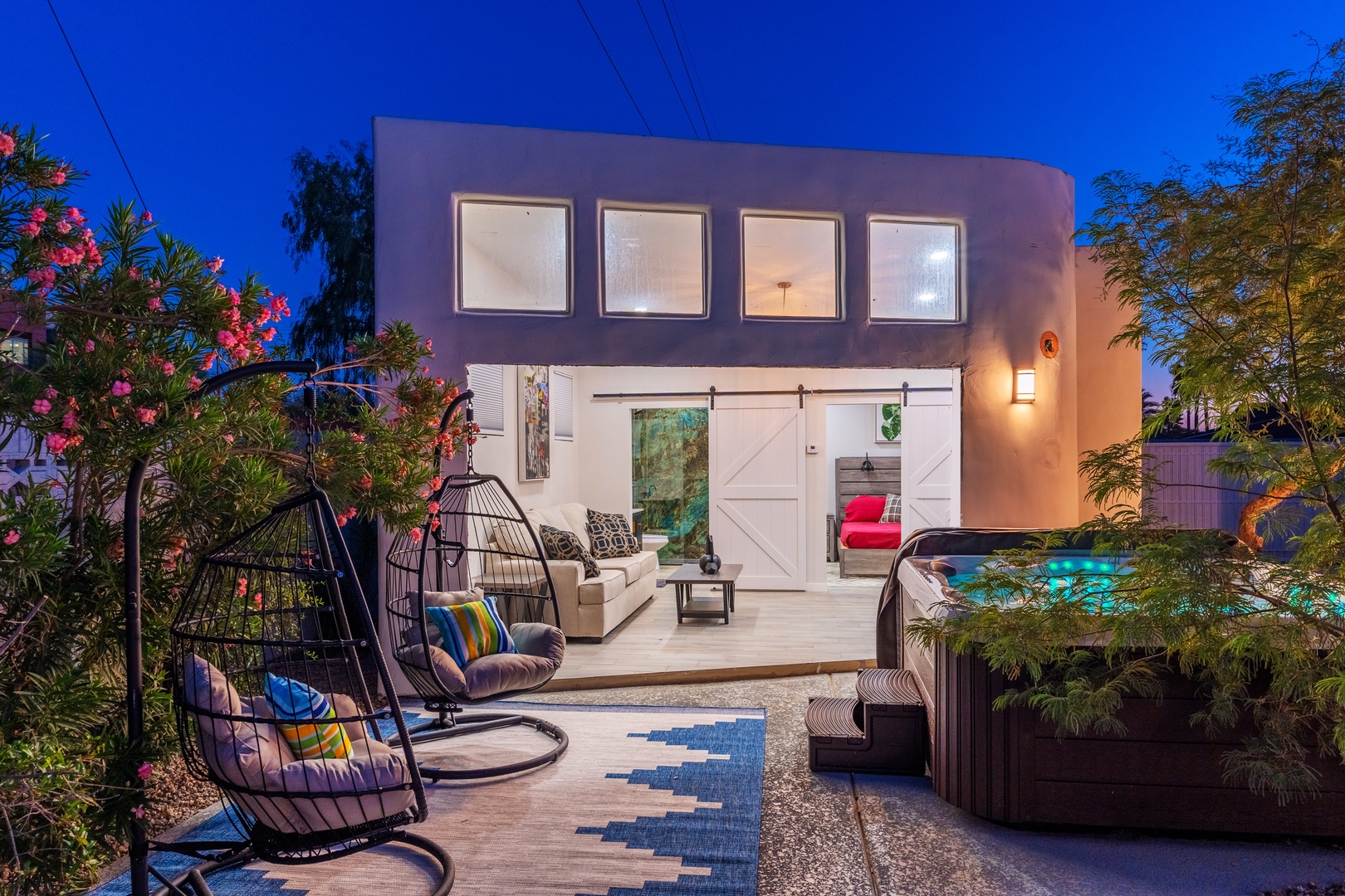 Modern desert retreat featuring a unique curved exterior, illuminated outdoor living space with sliding barn doors, hot tub, cozy swing chairs, and lush landscaping under vibrant evening skies.