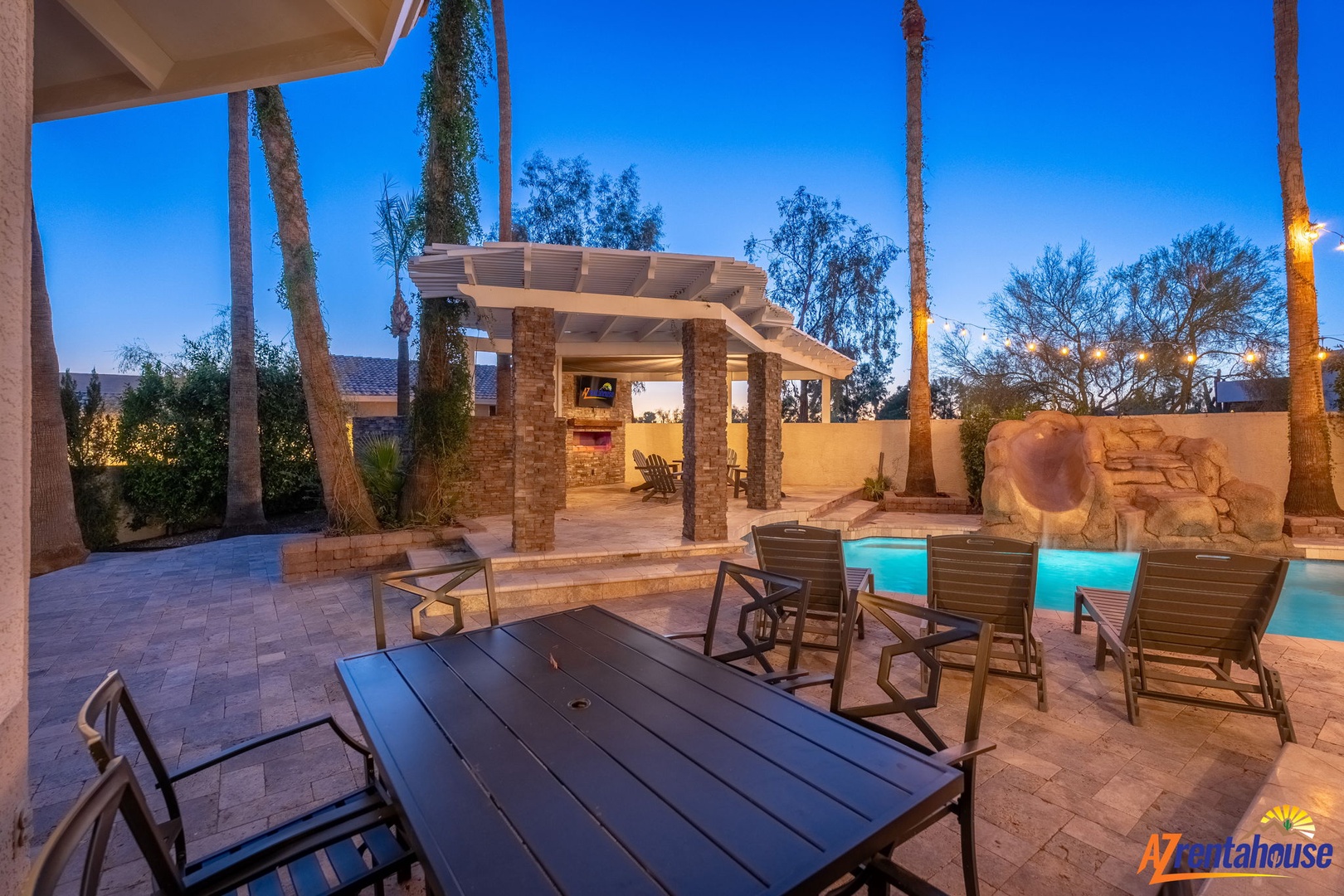 A resort-style backyard features an illuminated pool, outdoor kitchen with pergola, and palm trees framed by a vibrant twilight sky—perfect for evening gatherings.