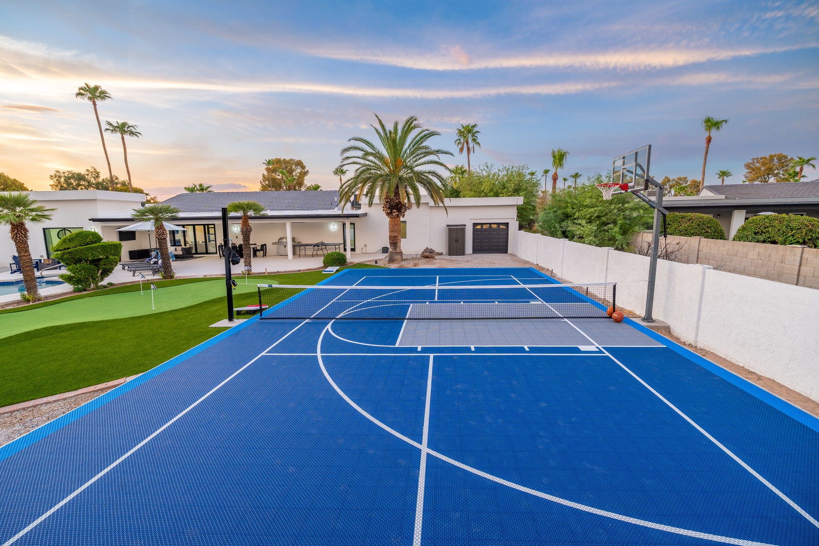 A modern desert oasis featuring a private multi-sport court perfect for basketball, tennis, and volleyball beneath swaying palms and stunning sunset skies. Challenge friends to a game or practice your serve in this luxurious backyard retreat.