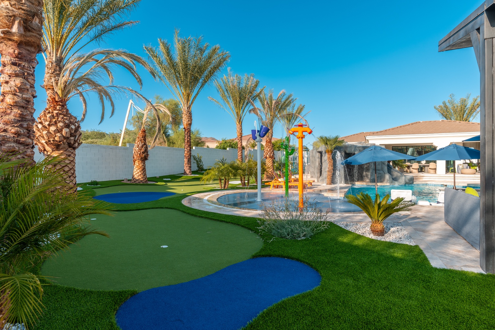 A private resort-style backyard with a sparkling pool, splash pad, putting green, and swaying palms set against endless Arizona blue skies.
