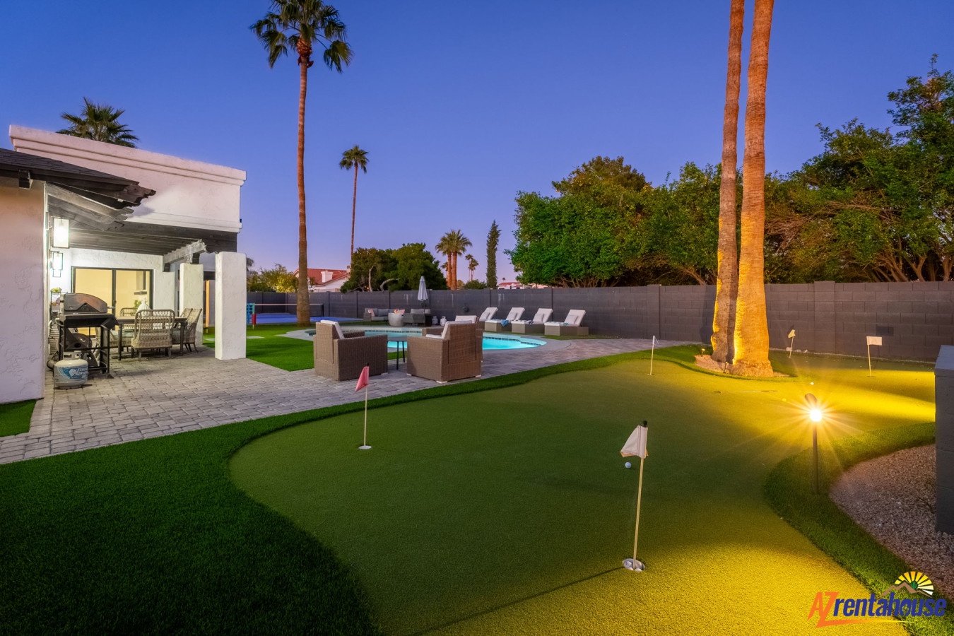 Stunning desert oasis backyard featuring a private putting green, sparkling pool, and modern patio under towering palm trees and evening lights.