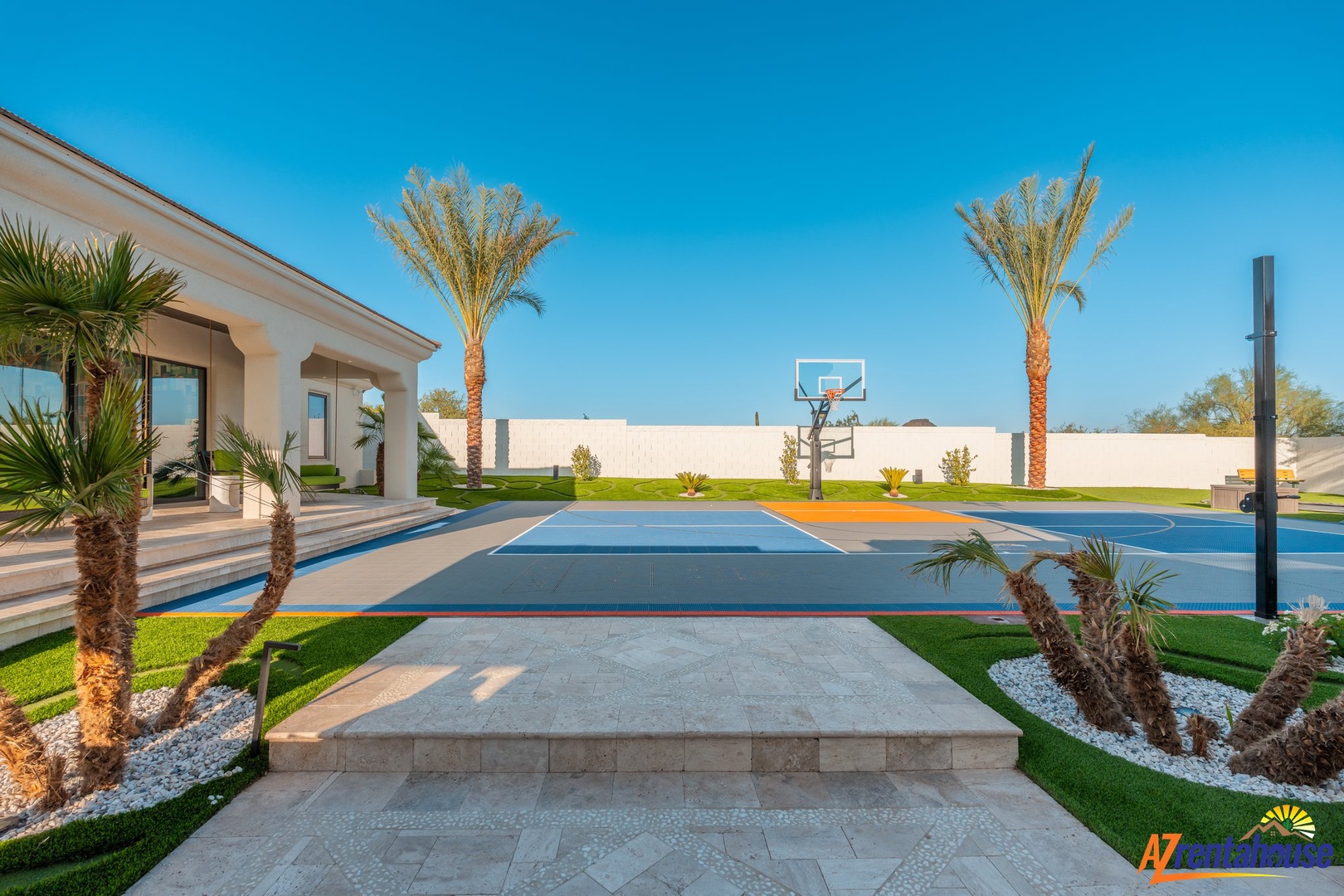 Modern luxury villa featuring a full basketball court surrounded by palm trees and pristine landscaping under clear blue skies.