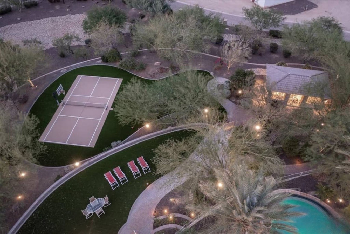 Desert estate with private tennis court, lush landscaping, illuminated pathways, and sparkling pool surrounded by natural Arizona terrain and mountain views.