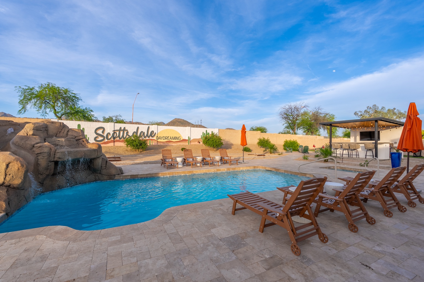 A stunning pool area features natural rock waterfall accents, comfortable loungers, and a shaded outdoor kitchen—perfect for sunny days and desert evenings under the open sky.