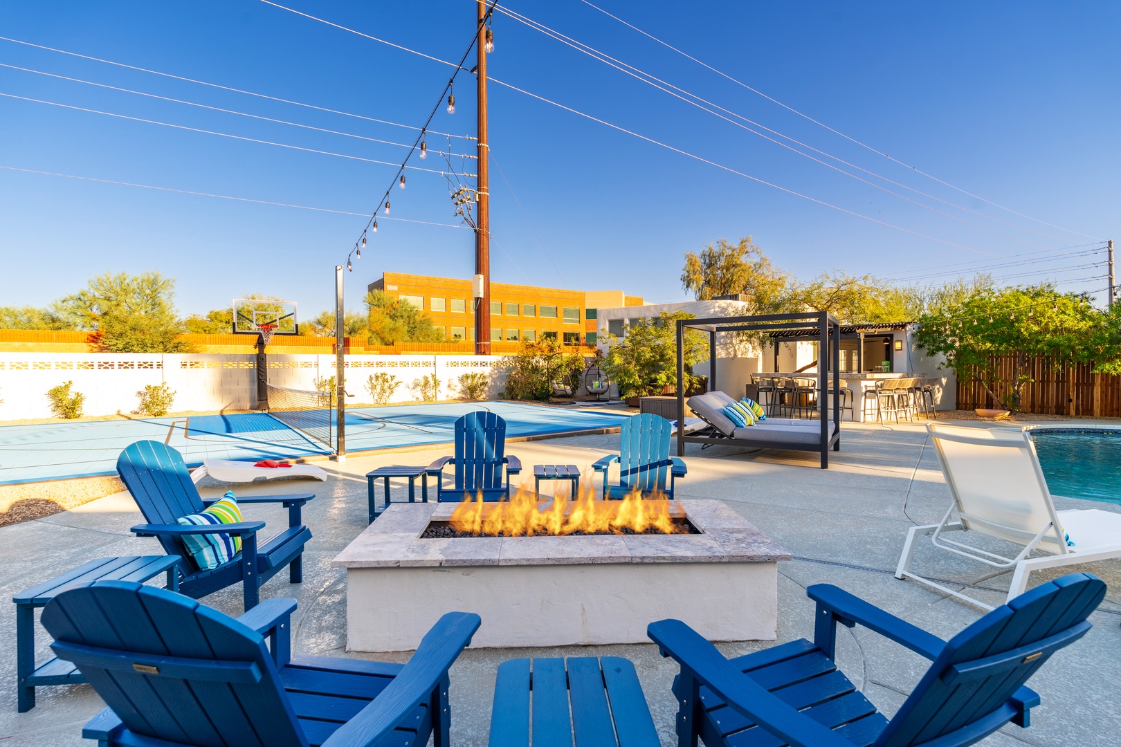 Gather around the outdoor fire pit surrounded by inviting blue Adirondack chairs, with a basketball court, pool, and shaded lounge area nearby—perfect for relaxing or staying active during your stay.