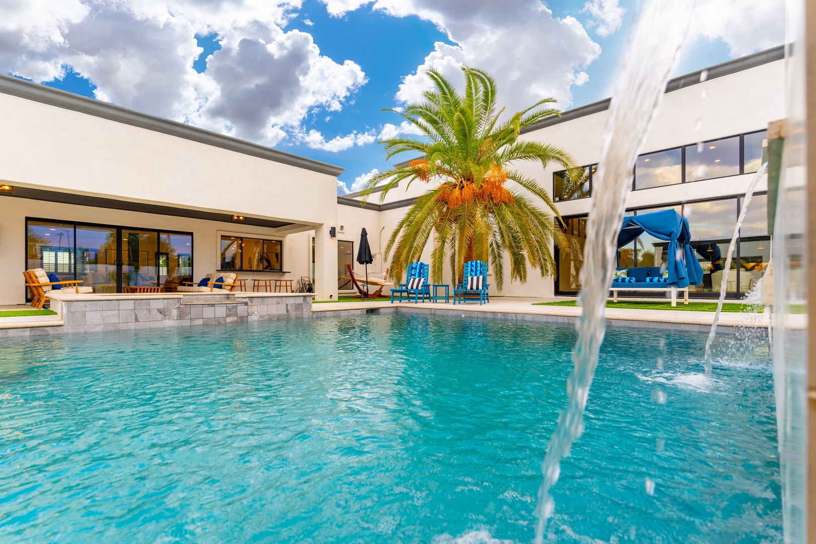 The property features a pristine swimming pool surrounded by modern architecture, vibrant palm trees, and comfortable poolside lounging areas—an ideal tropical retreat setting.