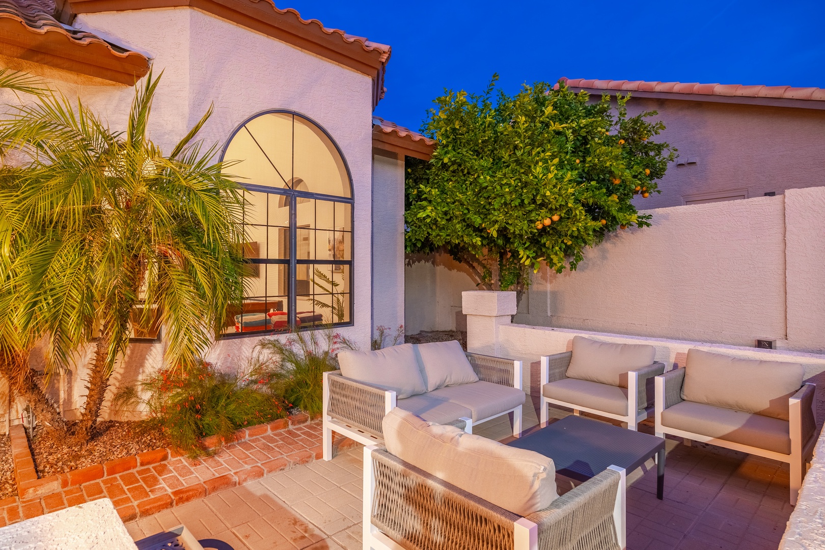 A Mediterranean-style villa showcasing warm stucco walls, terracotta roof tiles, and a distinctive arched window that frames the evening ambiance. Palm trees and citrus greenery accent the brick-paved courtyard.