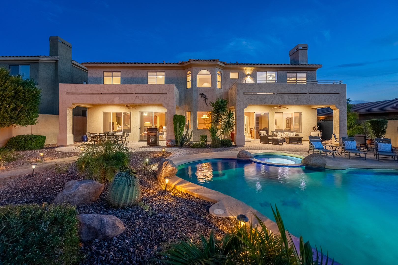 This stunning property showcases elegant desert landscaping, a sparkling pool with spa, and covered outdoor seating areas illuminated by warm ambient lighting under a twilight sky.