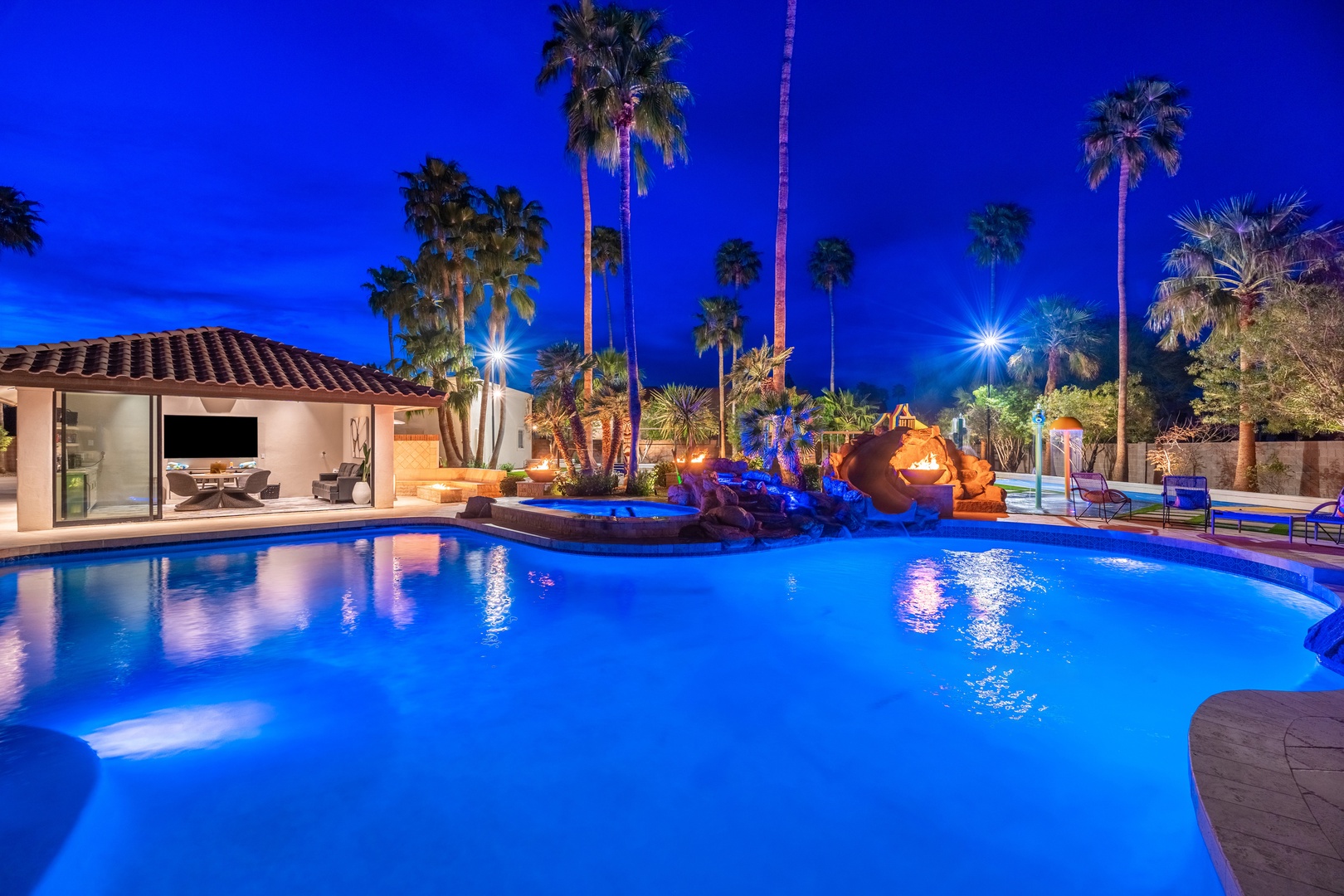 This stunning resort-style pool features illuminated waters surrounded by swaying palm trees and desert landscaping, creating an oasis atmosphere perfect for evening relaxation under the stars.