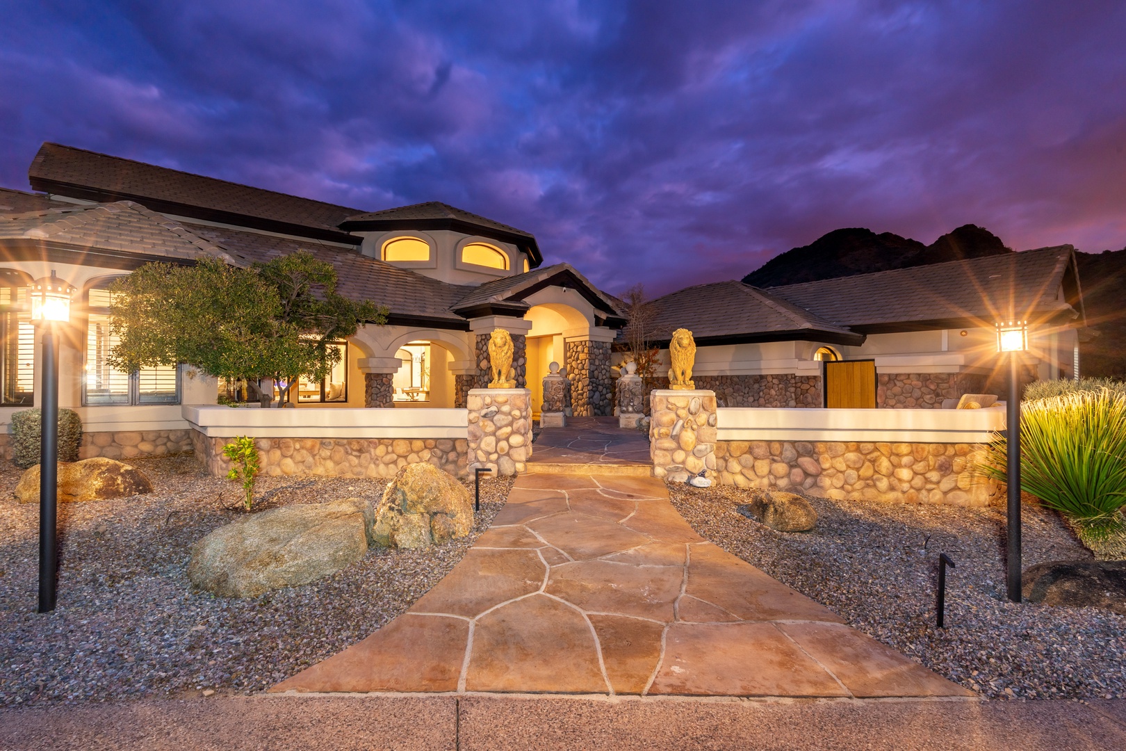 This grand desert estate features dramatic architecture with illuminated arches and lion-guarded entry pillars set against mountain backdrop, offering an impressive arrival experience at twilight.