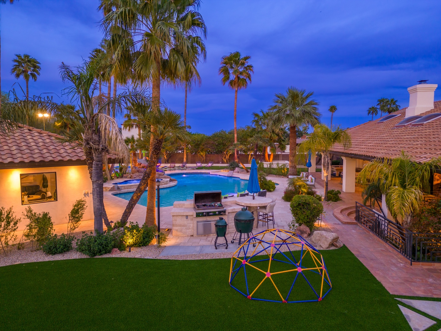 A twilight oasis featuring a sparkling pool, palm-lined patio, and vibrant play area—perfect for family fun and evening BBQs under desert skies.