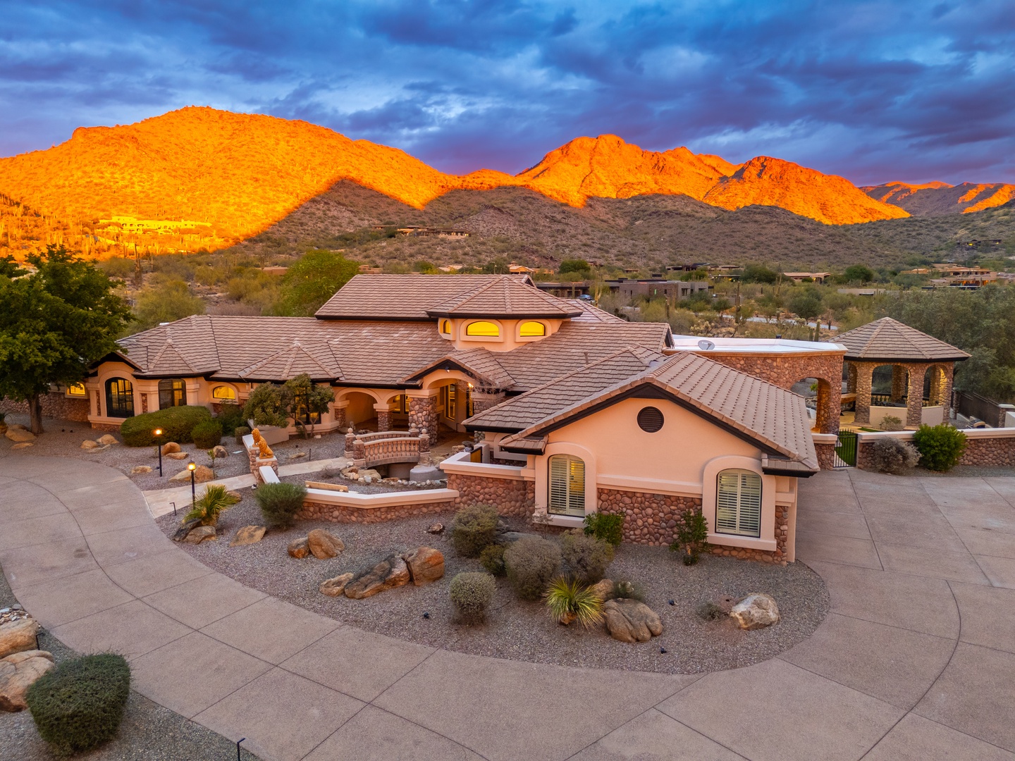 Spectacular desert estate framed by dramatic mountain peaks glowing in golden alpenglow, surrounded by natural landscaping and desert vegetation in a serene neighborhood setting.