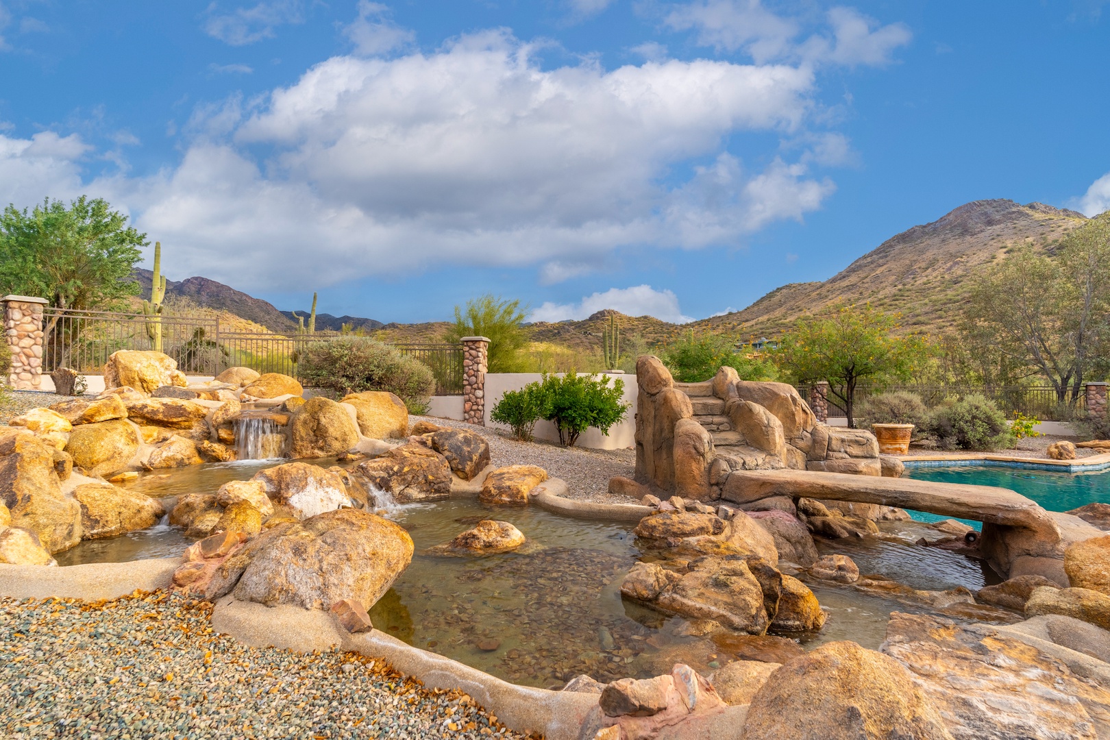 A serene desert oasis showcasing natural rock formations with flowing water features, set against stunning mountain views under an expansive blue sky with scattered clouds.