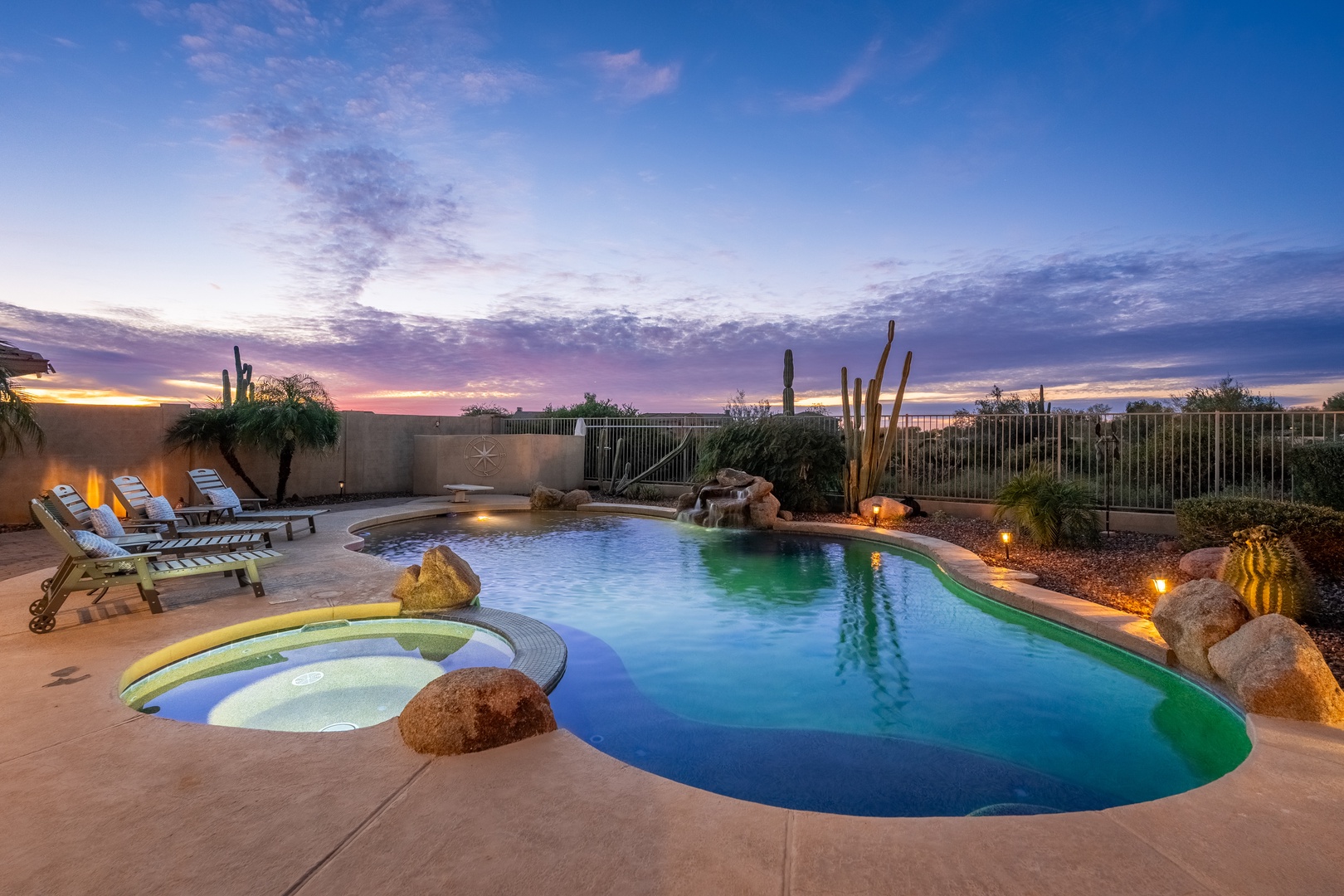 A stunning desert evening pool area glows under soft lighting, with comfortable loungers, hot tub, and natural rock features framed by cacti and mountain views beyond.