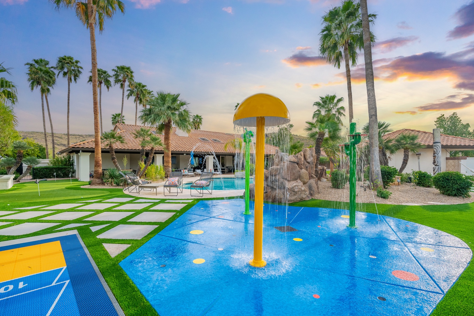 This stunning desert retreat features a vibrant splash pad and inviting pool area, surrounded by lush landscaping and swaying palms. The expansive lawn and dramatic sunset backdrop create a paradise perfect for families seeking relaxation and fun.