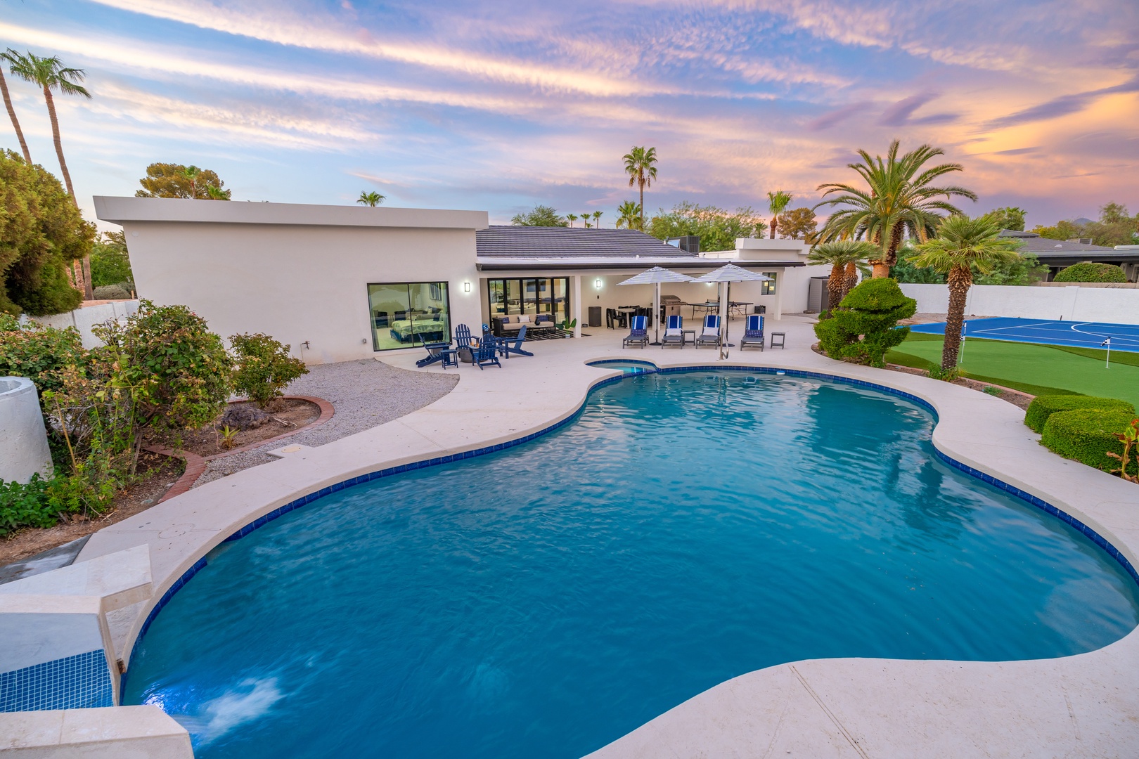 A striking aerial view of the modern property at dusk, featuring a large pool, manicured landscaping with palm trees, and a tennis court on the grounds—capturing the property's lush desert setting.