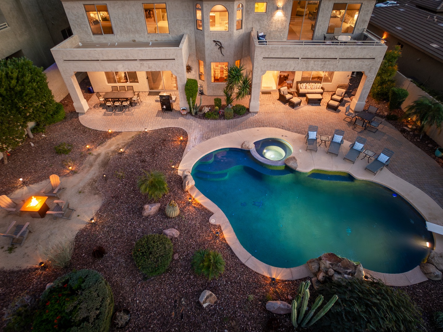 Aerial view captures the property's inviting outdoor living spaces, featuring a resort-style pool with integrated hot tub, covered patios with dining and lounge areas, and tasteful desert landscaping illuminated by ambient lighting.
