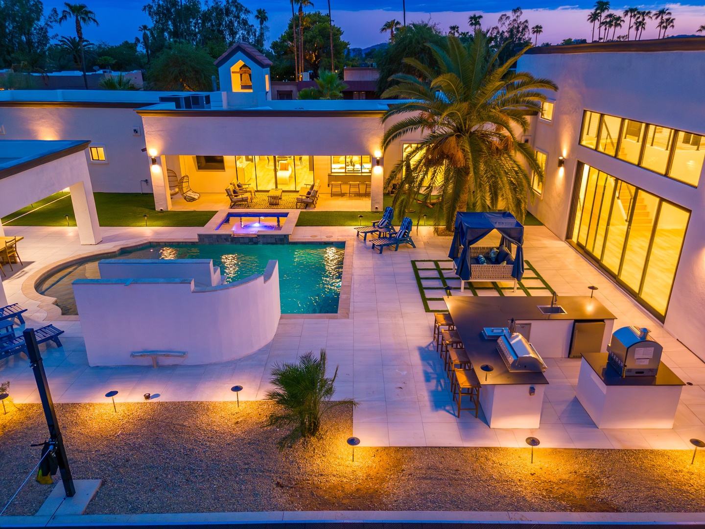 Modern desert retreat featuring a sparkling pool, expansive courtyard, and mountain backdrop surrounded by palms, captured at twilight as ambient lighting illuminates the property's contemporary architectural design.